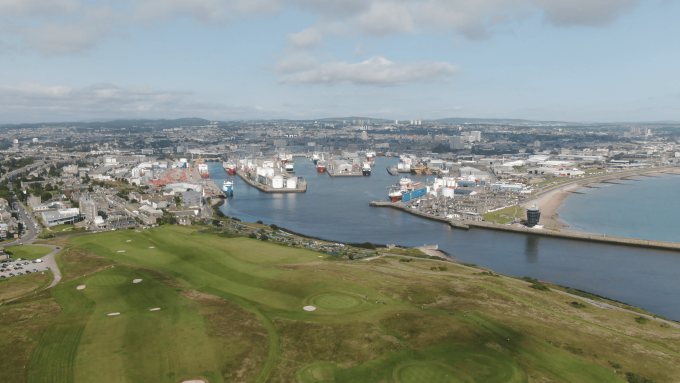 Aerial view of a coastal city featuring a large port with multiple ships, surrounded by urban buildings, green open fields, and a sandy beach along the shoreline.