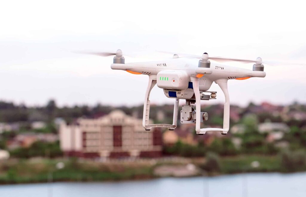 A white quadcopter drone hovers in the air with a blurred background of buildings and a river.