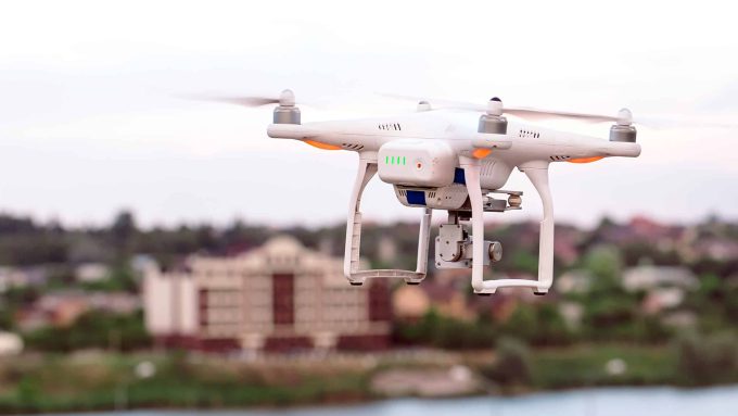 A white quadcopter drone hovers in the air with a blurred background of buildings and a river.