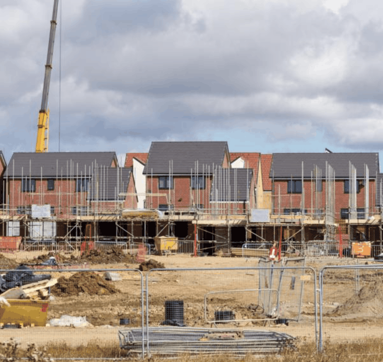 A row of partially constructed houses with scaffolding in place and construction materials scattered around, under a cloudy sky. A crane is visible in the background.