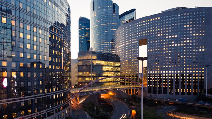 A view of modern high-rise buildings with illuminated windows in a cityscape during dusk. A bridge connects some of the buildings, and a curved road runs between them.