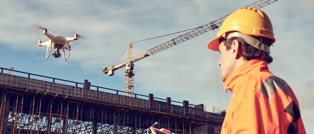 A construction worker in an orange outfit and helmet operates a drone at a construction site with a crane and building structure in the background.