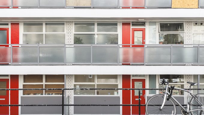 A modern apartment building with frosted glass balconies. Several red doors are visible, and a bicycle is parked at the front railing.