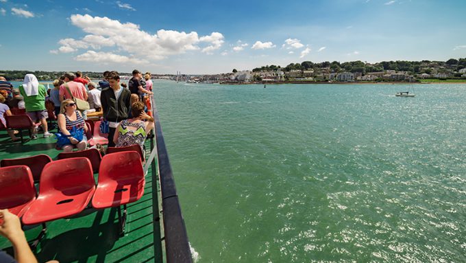 A group of people stand and sit on red seats on a boat, looking out at a coastal town and blue-green waters under a partly cloudy sky.