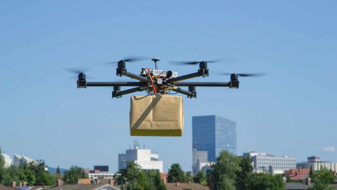 A drone carrying a package flies over a cityscape with buildings and trees visible in the background under a clear blue sky.