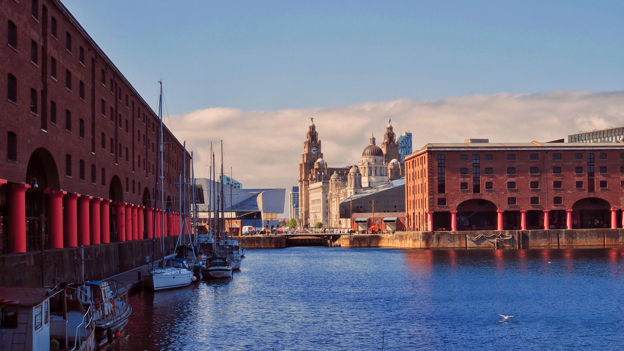A waterfront scene along the Mersey with docked boats, historic red brick buildings on both sides, and a view of an ornate cathedral in the distance under a partly cloudy sky.
