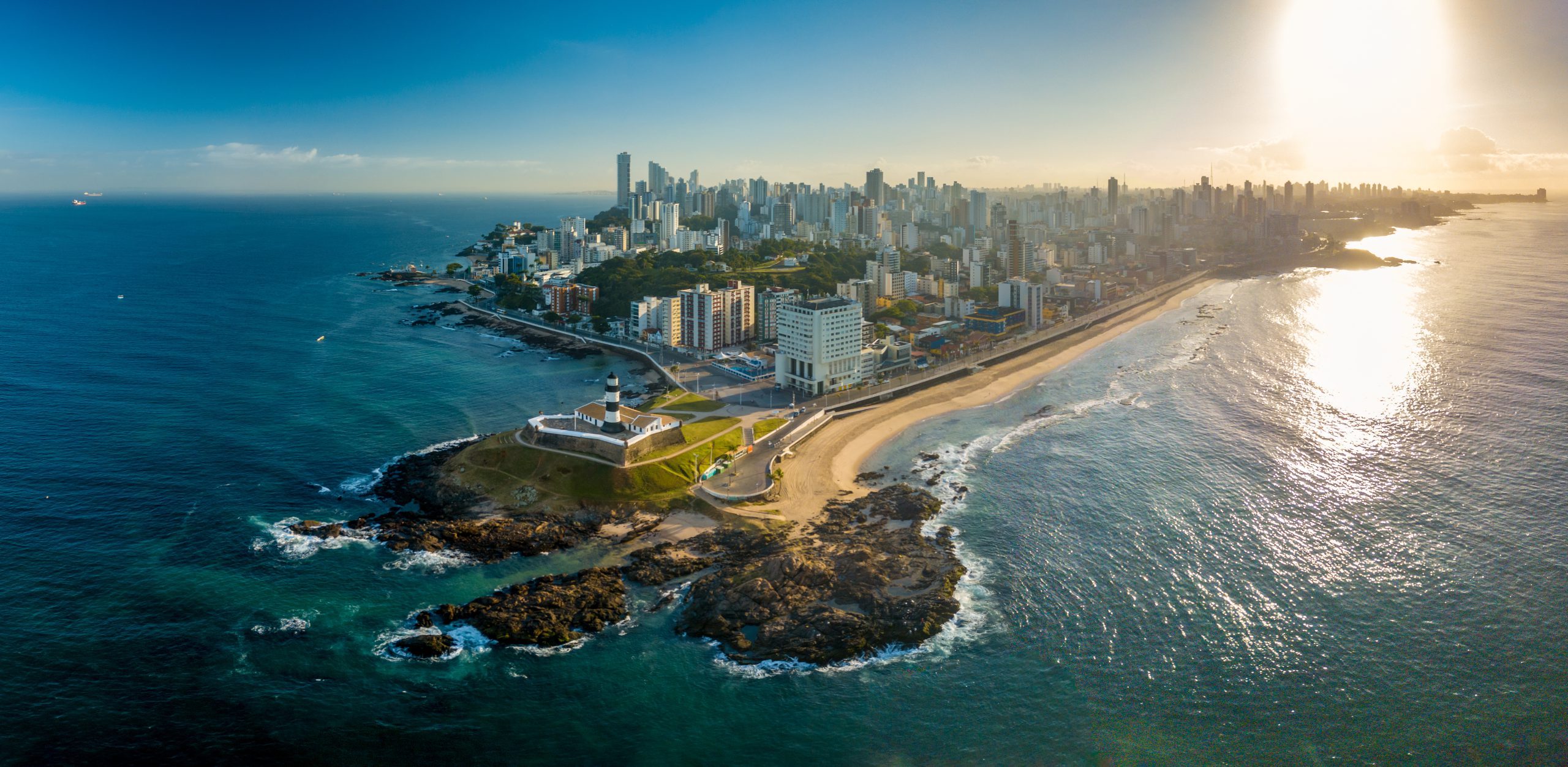 Aerial view of a coastal city with a mix of modern high-rise buildings and a historic lighthouse near the shoreline, surrounded by a sandy beach and the ocean under a clear sky with the sun setting, embodying the principles of resilient and connected cities.