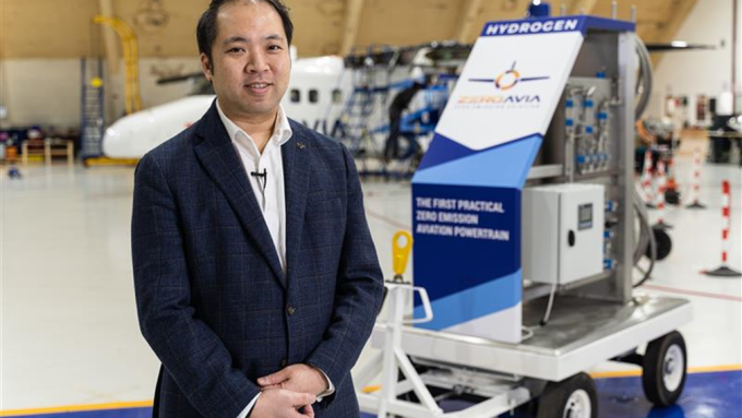 A man stands in an aircraft hangar near a display labeled "HYDROGEN" and "The First Practical Zero Emission Aviation Powertrain" by ZeroAvia, with an aircraft visible in the background.