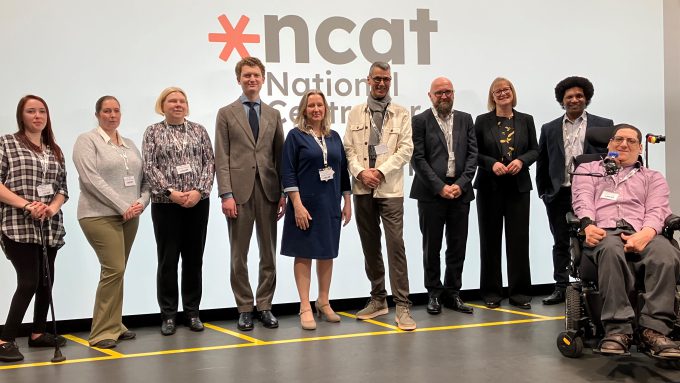Group of ten people posing for a photo on stage in front of a screen with the NCAT logo. One person is in a wheelchair. They are all dressed in business or casual attire.