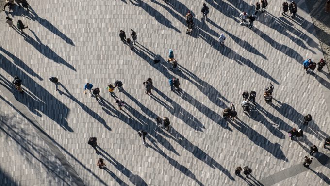 Aerial view of people walking and standing on a large tiled plaza, casting long shadows in the sunlight.