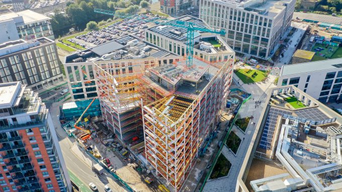 An aerial view of a partially constructed high-rise building, surrounded by other modern buildings in an urban area, showcases the dynamic interplay of innovation and procurement. Construction equipment and materials are visible at the site.