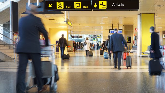 Passengers with luggage walk through an airport terminal beneath signs indicating directions for departures, check-in, taxi, bus, and arrivals.
