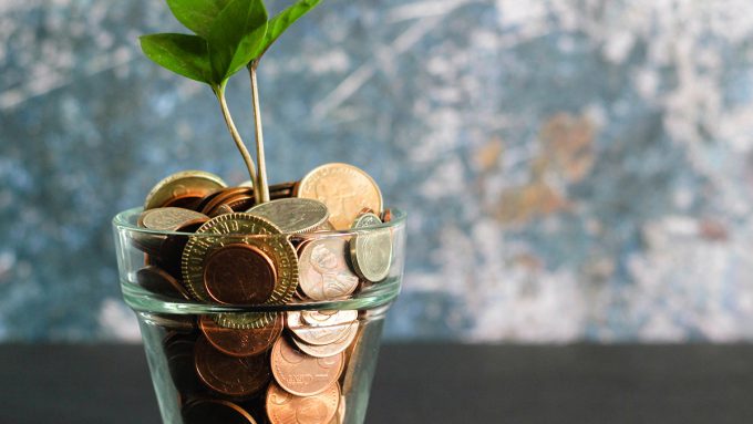A small green plant growing from a glass container filled with a mix of coins against a textured background.