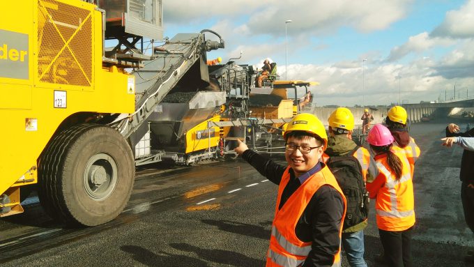 Workers in high-visibility vests and hard hats oversee road construction machinery on a sunny day. One worker points towards the machinery, highlighting the ongoing paving process using sustainable road tech.