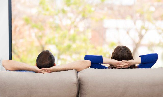 Two people are sitting on a couch with their backs to the camera. They are leaning back, resting their heads on their hands, and looking out at a blurred outdoor scene through a large window.