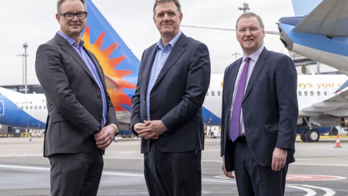 Three men in business attire stand on an airport tarmac with two commercial airplanes in the background.