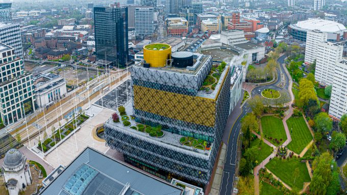 Aerial view of a cityscape featuring a modern building with a landscaped rooftop, surrounded by roads, greenery, and other buildings on a cloudy day in Birmingham, highlighting the city's tech advancements fueled by recent funding.