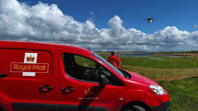 A Royal Mail van is parked on grass next to a safety net fence. A person in an orange jacket is operating a drone, as part of a drone mail delivery test, in a cloud-filled sky over the grassy field.