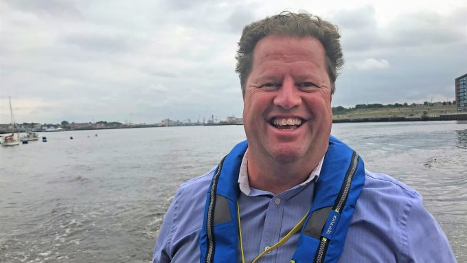 A man smiling at the camera wearing a blue life vest, with a cloudy sky and waterfront in the background, hints at the port's maritime decarbonisation efforts.