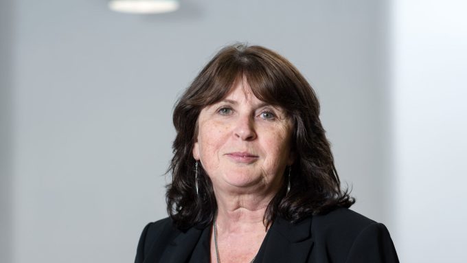 A woman with shoulder-length dark hair, wearing a black blazer and necklace, is standing in front of a plain background with a light fixture above her.