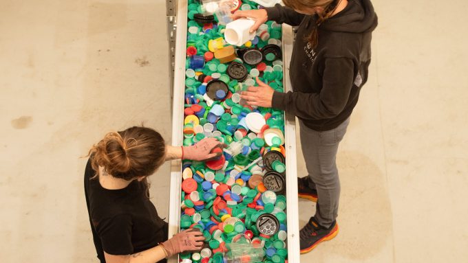 Two people sorting various colorful plastic bottle caps on a conveyor belt.