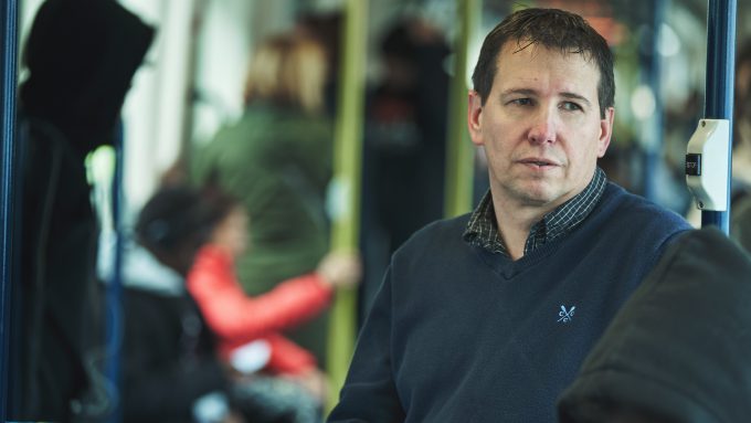A man in a blue sweater sits on a bus, looking to his left. Other passengers, some in hooded jackets, are seated and standing in the background, all mindful of transport safety.