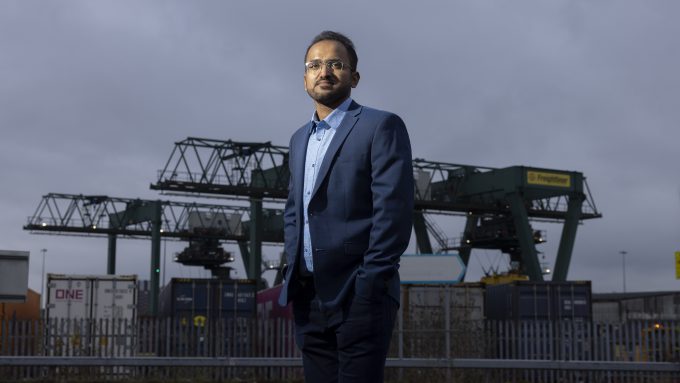 A man in a blue suit and glasses stands in front of industrial cranes and shipping containers under a cloudy sky.