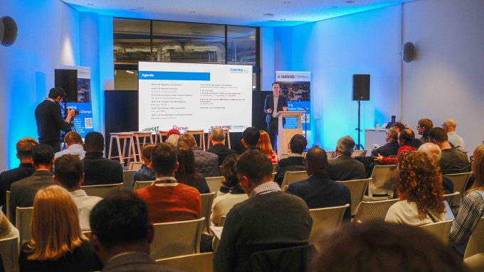 A group of people seated and facing a speaker and a presentation screen in a conference room. The screen displays an agenda for the Clean Futures Catalyst while the speaker presents and another person stands nearby.