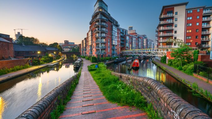 A peaceful canal scene in the West Midlands at dusk, featuring a narrow brick towpath leading between two canals. Modern red-brick apartment buildings line the water, with balconies overlooking the moored narrowboats. A footbridge crosses the canal, and soft lighting reflects on the water. The sky is clear with a warm evening glow.