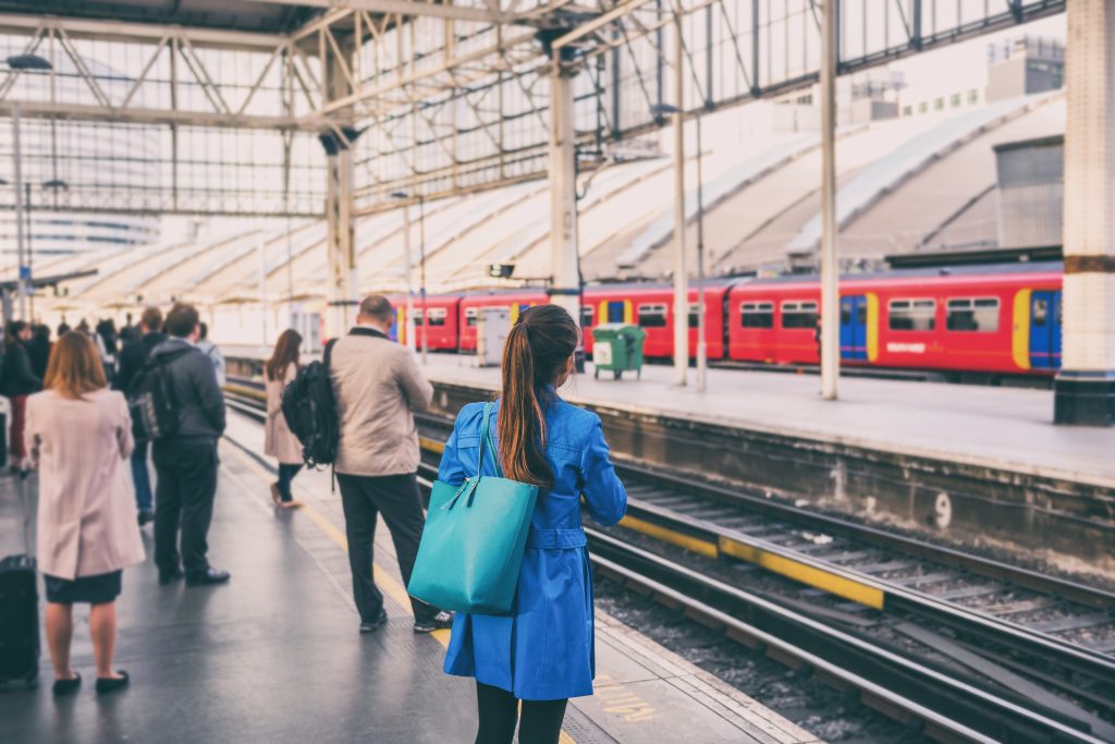 A group of people stand on a train platform during morning rush hour, dressed in business attire and waiting for a commuter train. The scene is slightly blurred to show motion and the hustle of daily travel.
