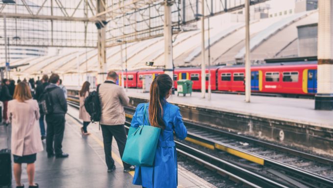 A group of people stand on a train platform during morning rush hour, dressed in business attire and waiting for a commuter train. The scene is slightly blurred to show motion and the hustle of daily travel.