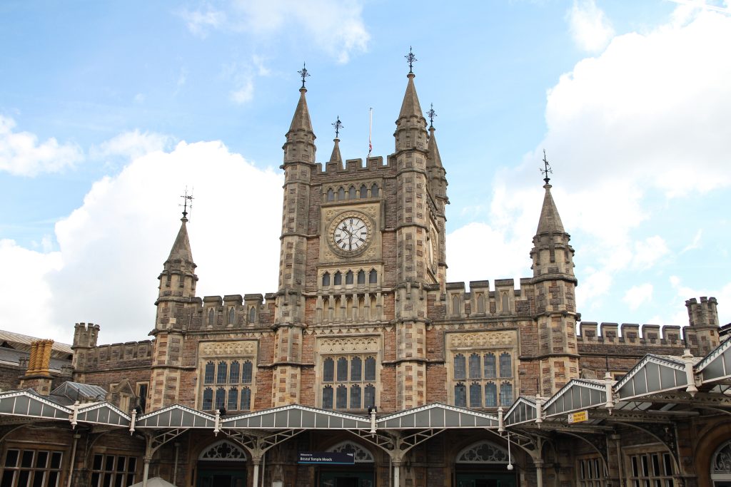 The historic facade of Bristol Temple Meads railway station, featuring a large central clock tower with four pointed spires and a mix of Gothic-style stonework and arched windows. A canopy supported by decorative ironwork extends across the station entrance, and the sky is partly cloudy with patches of blue.