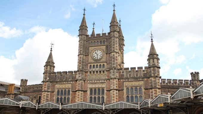 The historic facade of Bristol Temple Meads railway station, featuring a large central clock tower with four pointed spires and a mix of Gothic-style stonework and arched windows. A canopy supported by decorative ironwork extends across the station entrance, and the sky is partly cloudy with patches of blue.